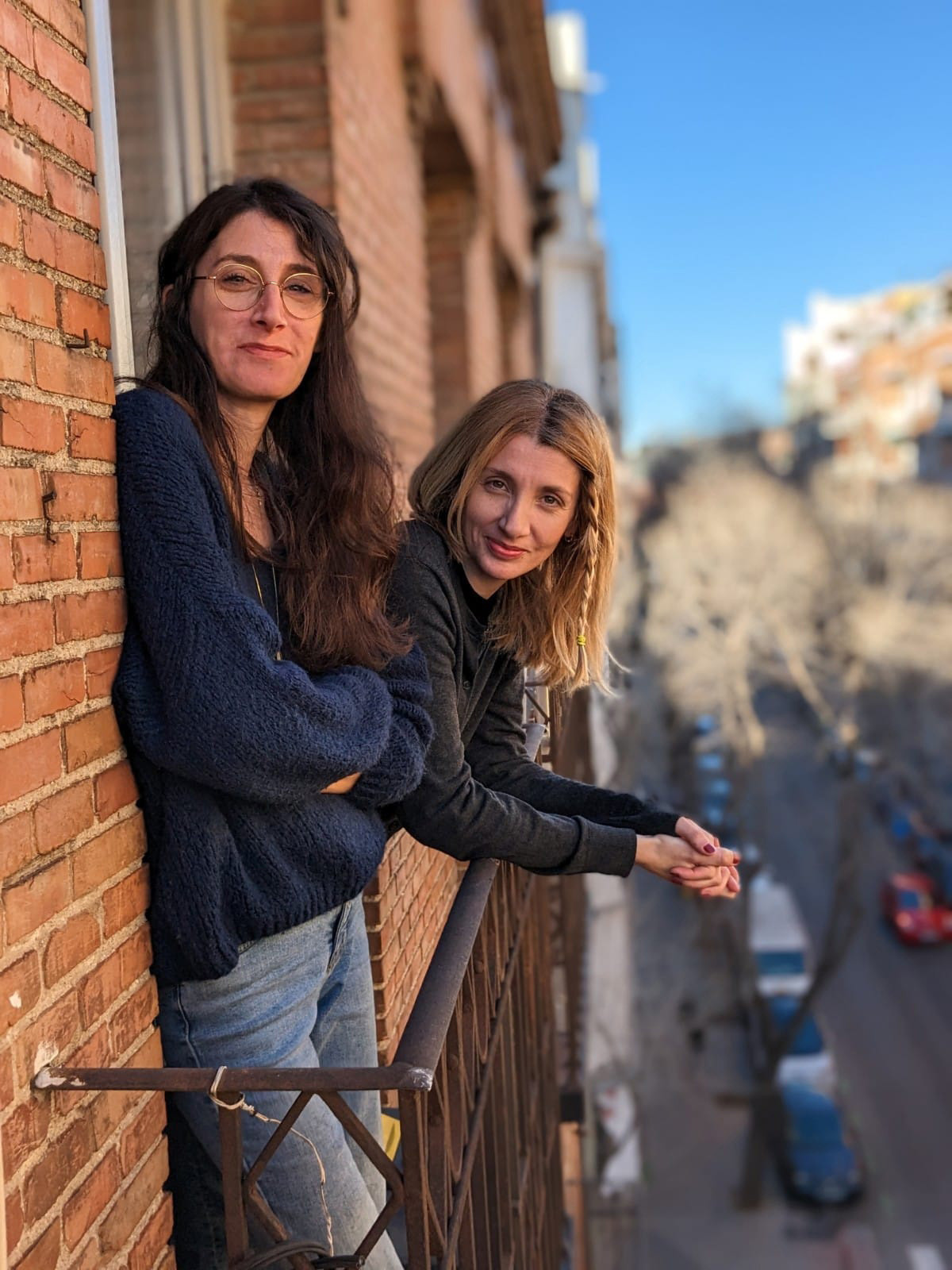 Two white women with long hair leaning against a brick building wearing comfortable sweaters