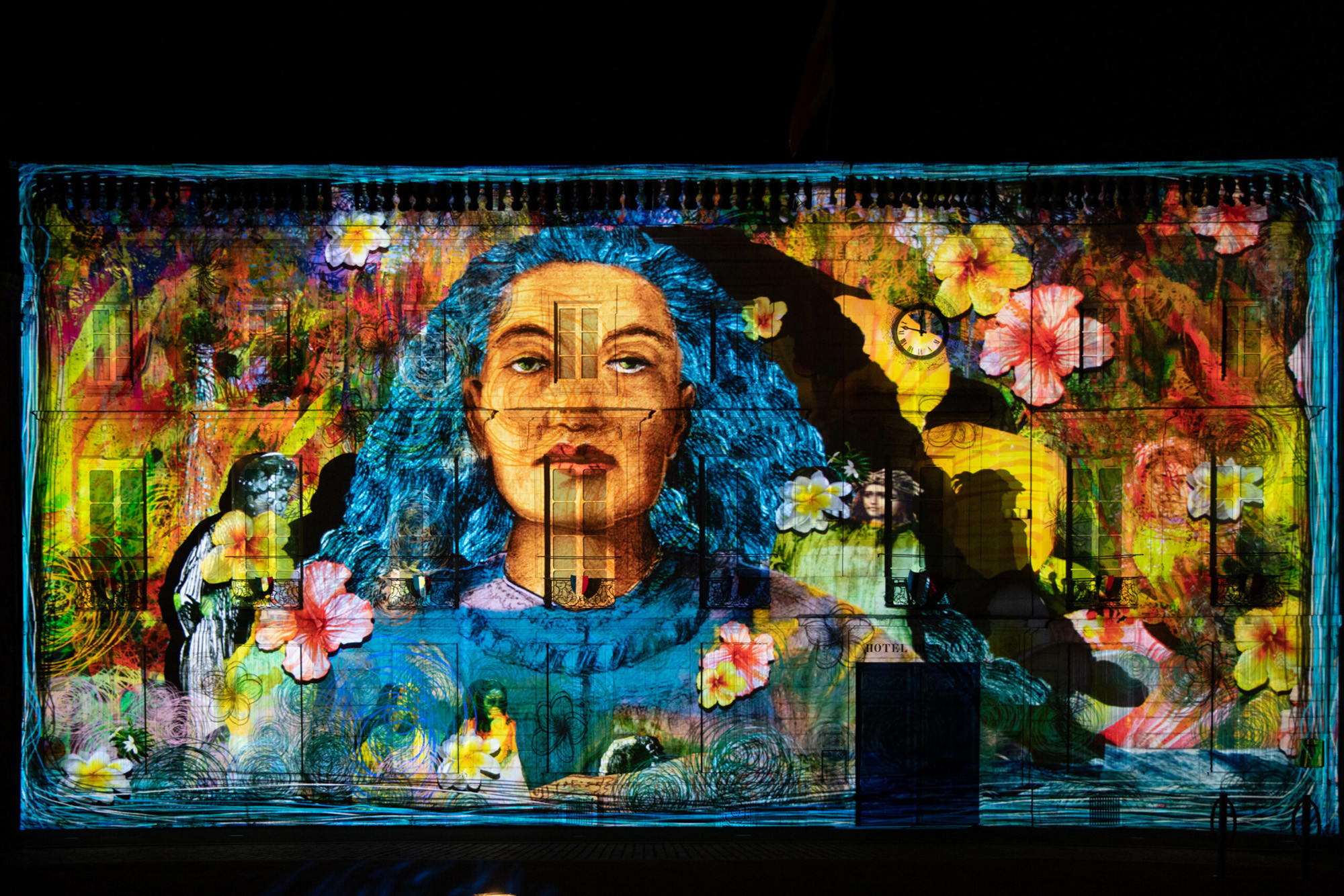 Nighttime photo of a flat building front with images of a blue-haired woman, flowers and other motifs projected on it.